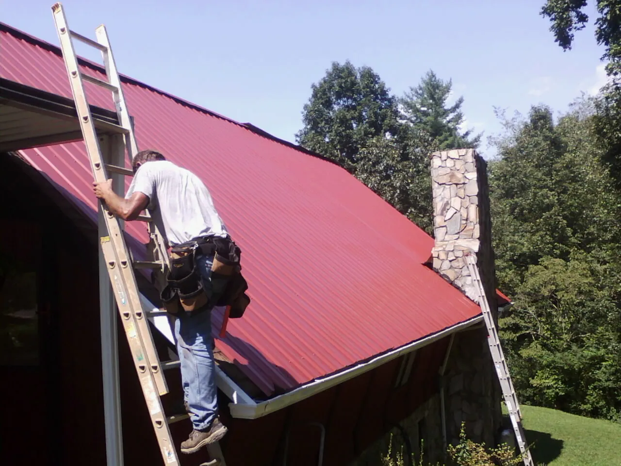 Roofer on a ladder beside a red metal roof and stone chimney — Soffit & Fascia Repair by Roofing Brothers in Reardan